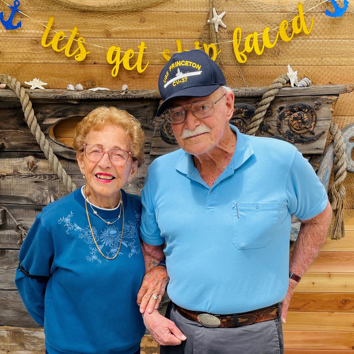 An elderly couple smiling and posing together indoors against a decorative backdrop.