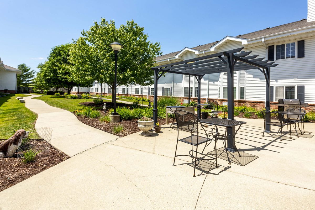 A sunny outdoor patio area with a pergola, tables and chairs, surrounded by well-maintained landscaping.
