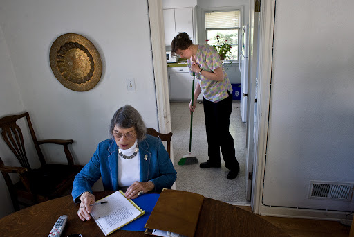 An elderly woman sits at a table while a young man cleans the floor in a cozy home environment.