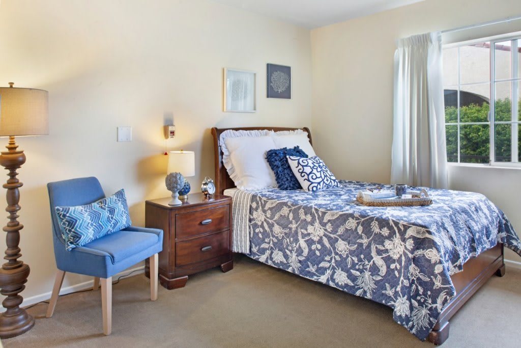 A cozy bedroom featuring a floral-patterned bedspread, a bedside table with a lamp, and a comfortable chair in the corner.