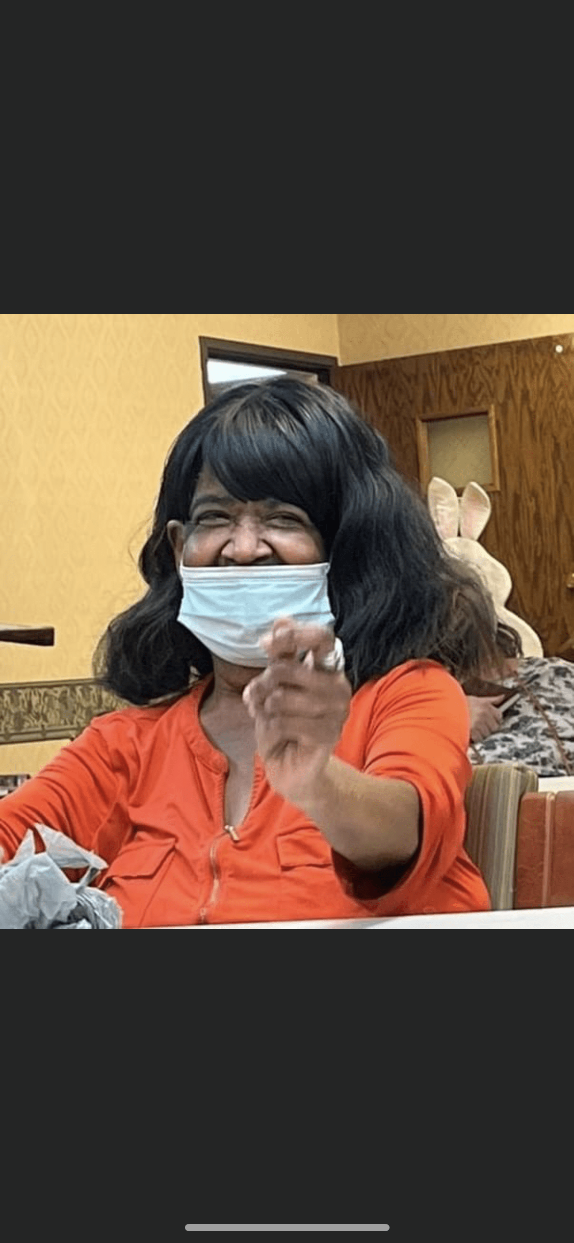 A woman wearing an orange shirt and mask smiles indoors.