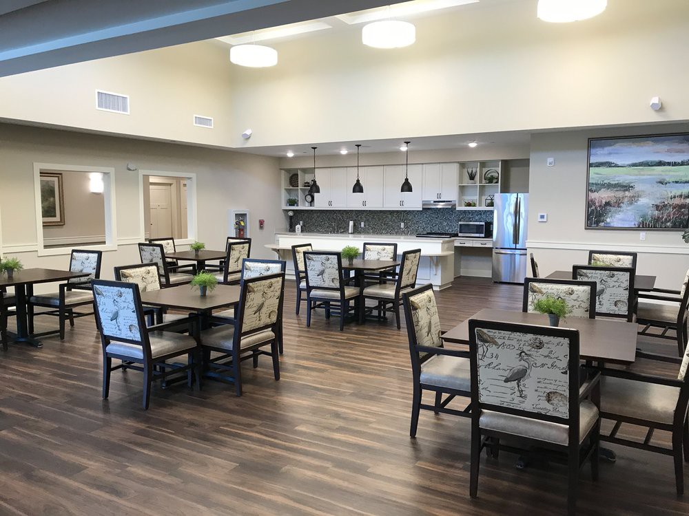 A modern dining area with a kitchen, featuring tables and chairs arranged on polished wooden flooring.