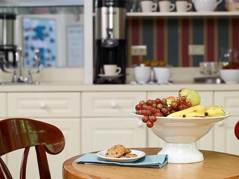A cozy kitchen setting with a fruit bowl and cookies on a table, accompanied by a coffee maker in the background.
