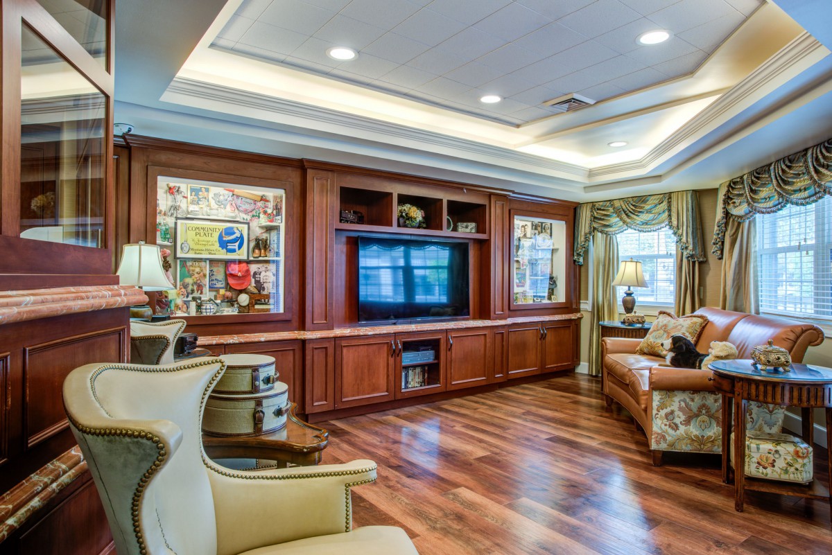 A spacious living room featuring hardwood flooring, a large television set into a wooden entertainment center, comfortable sofas, and decorative wall art under ceiling lights.