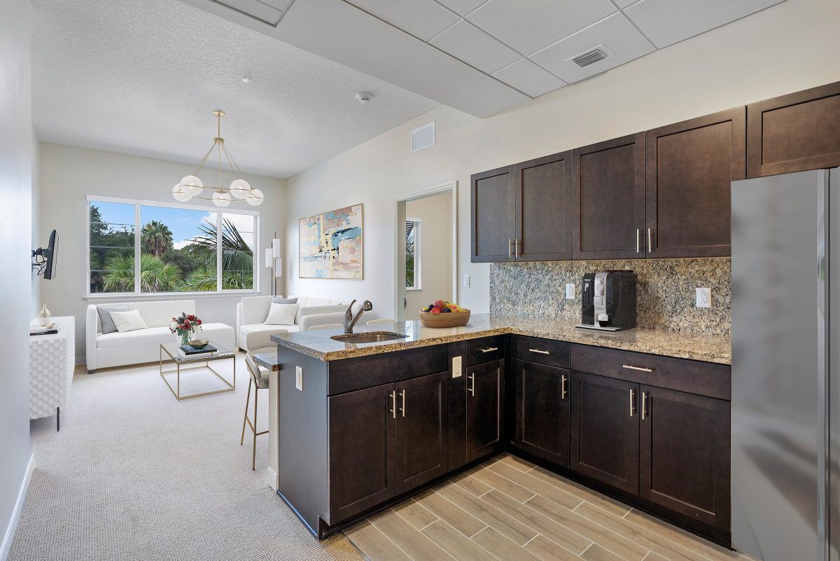 A modern kitchen with dark cabinets and a connected dining area, featuring ample natural light from the large window.