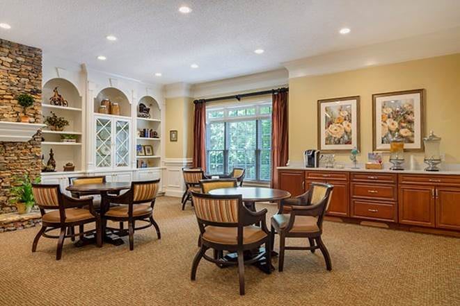 A cozy living room with tables, chairs, bookshelves, and large windows that let in natural light.