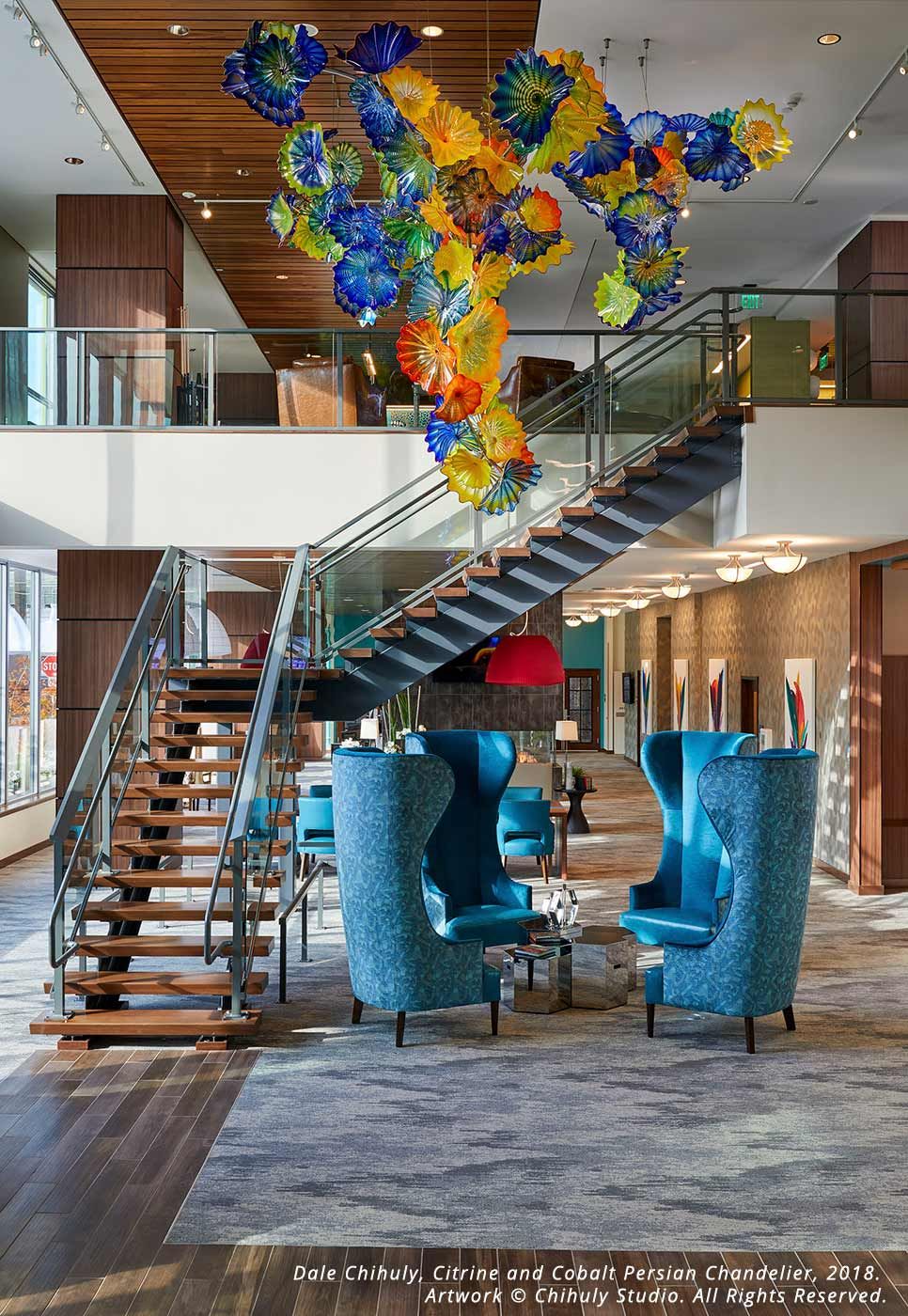 A modern lobby featuring a striking staircase and colorful hanging art, complemented by blue high-back chairs in the seating area.