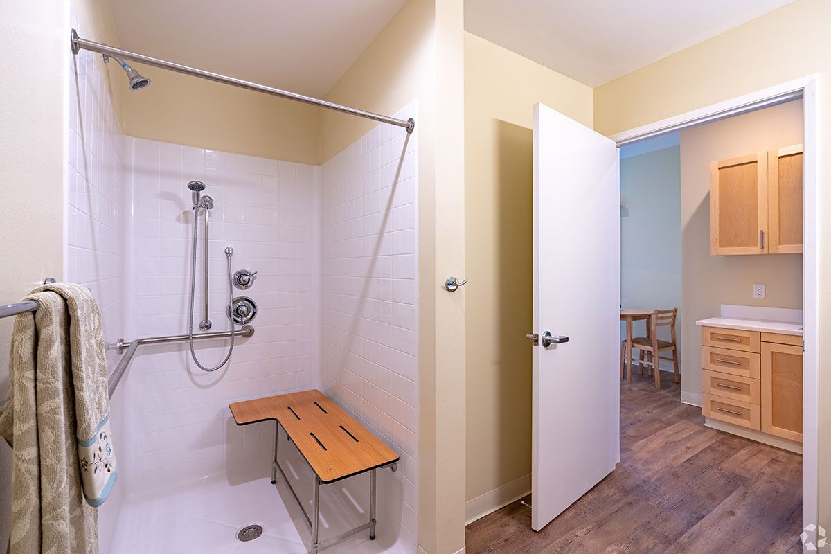 An accessible bathroom featuring a shower with a safety rail and bench seat, adjacent to an open door leading to another room.