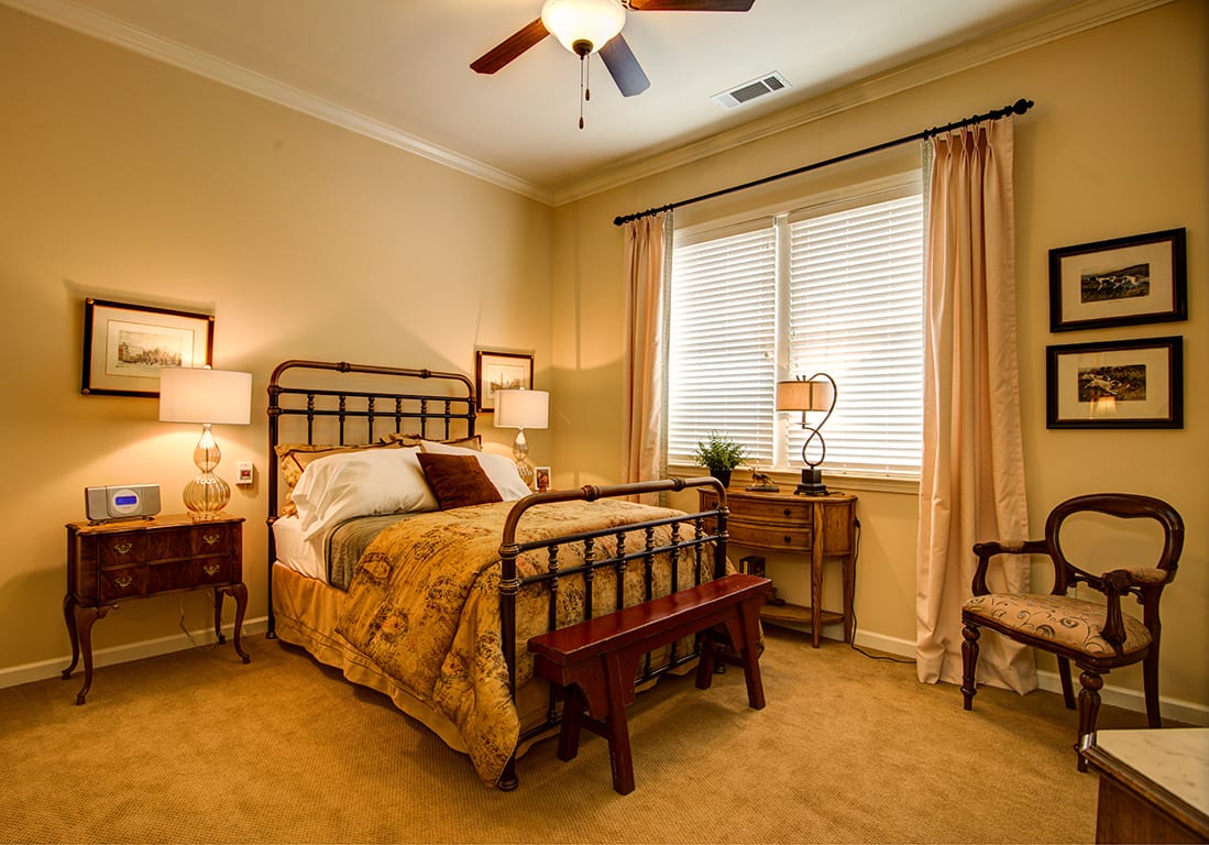 A cozy bedroom with warm lighting, featuring a bed with decorative pillows, bedside tables with lamps, and large windows with curtains.