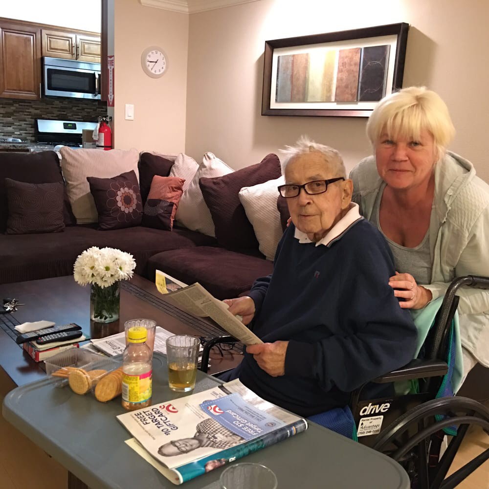 An elderly person sitting in a wheelchair reading a magazine, accompanied by another person in a cozy living room.