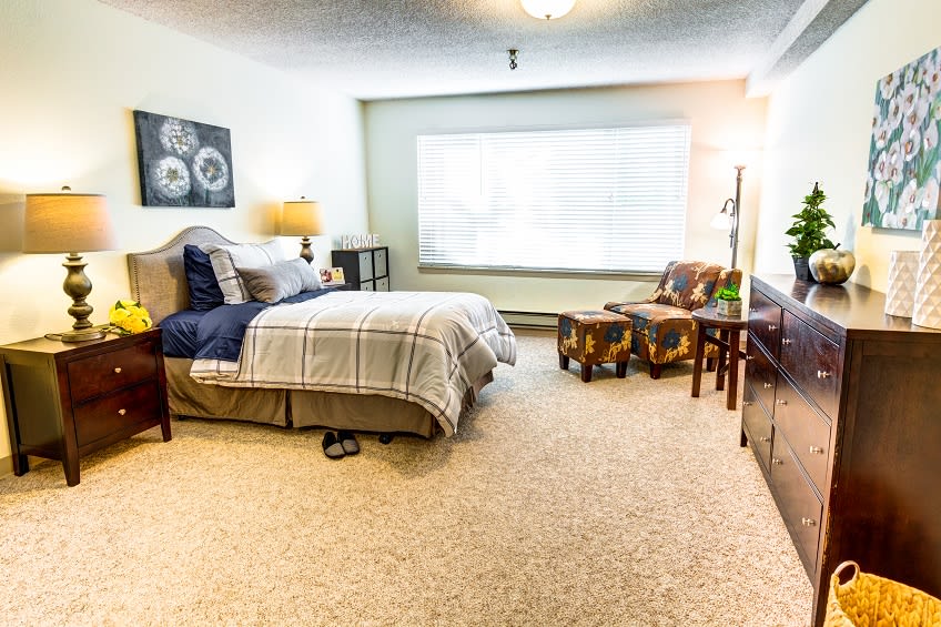 A cozy bedroom with a bed, bedside lamp, window seating area, and carpeted floor.