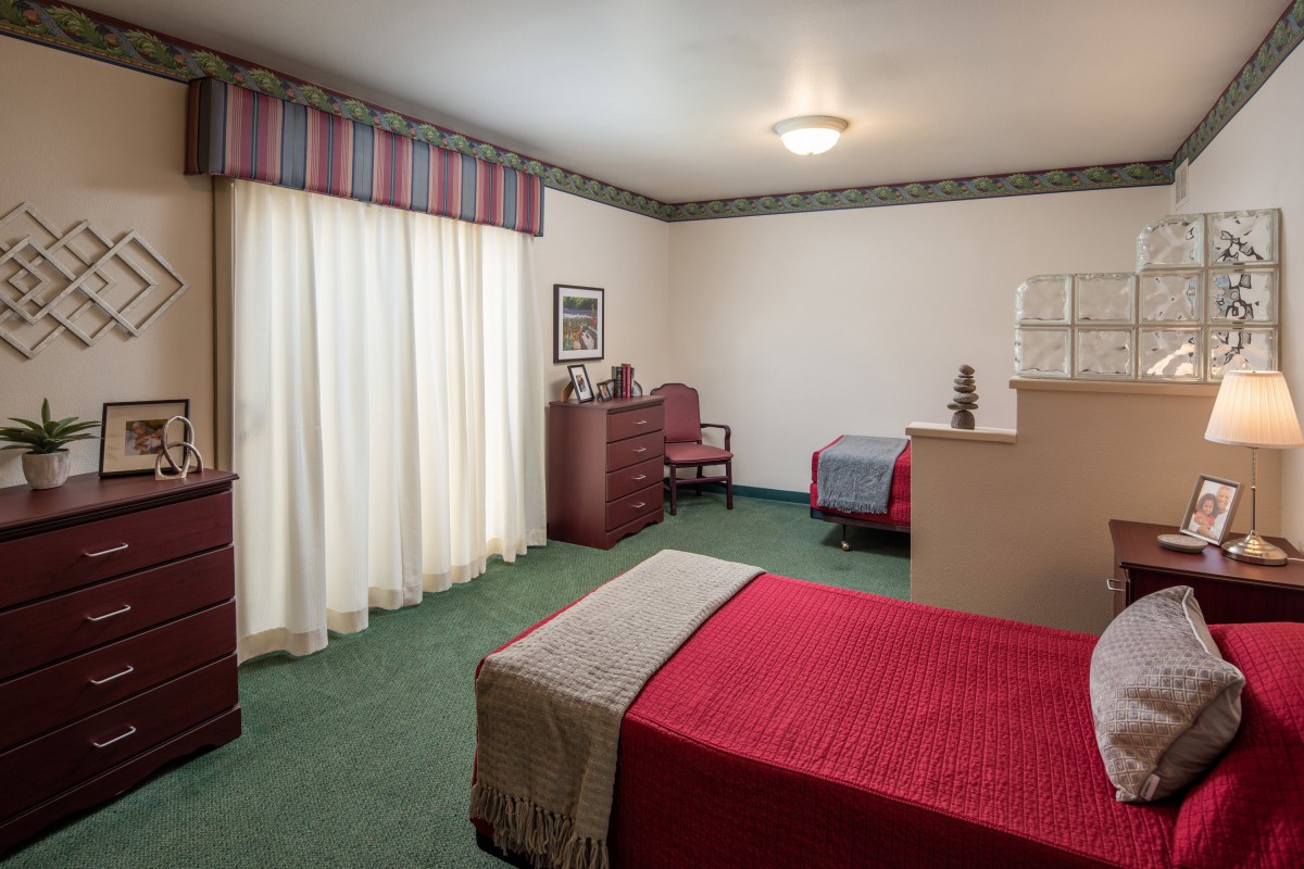 A simple bedroom with a red bedspread, dresser, and carpeted floor.