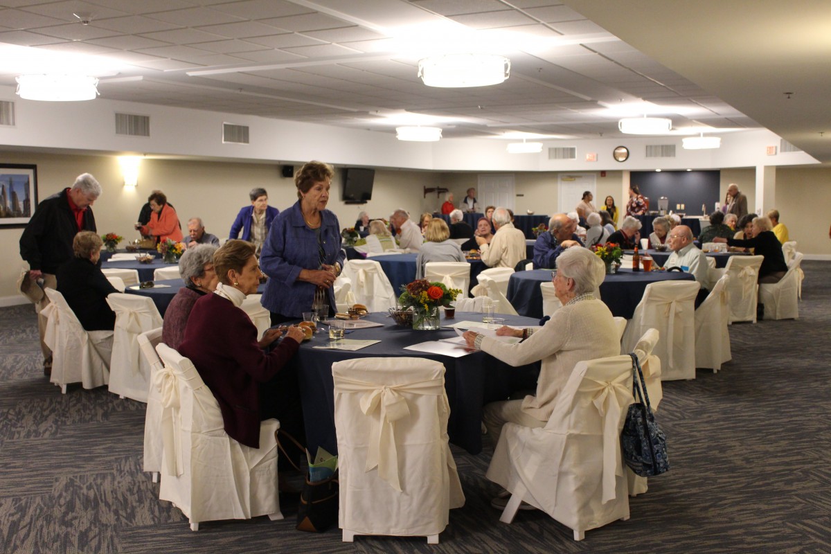 A community gathering with seniors seated at tables in a large indoor venue.