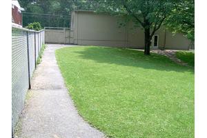 A grassy yard with a pathway, fenced area, tree, and building in the background.