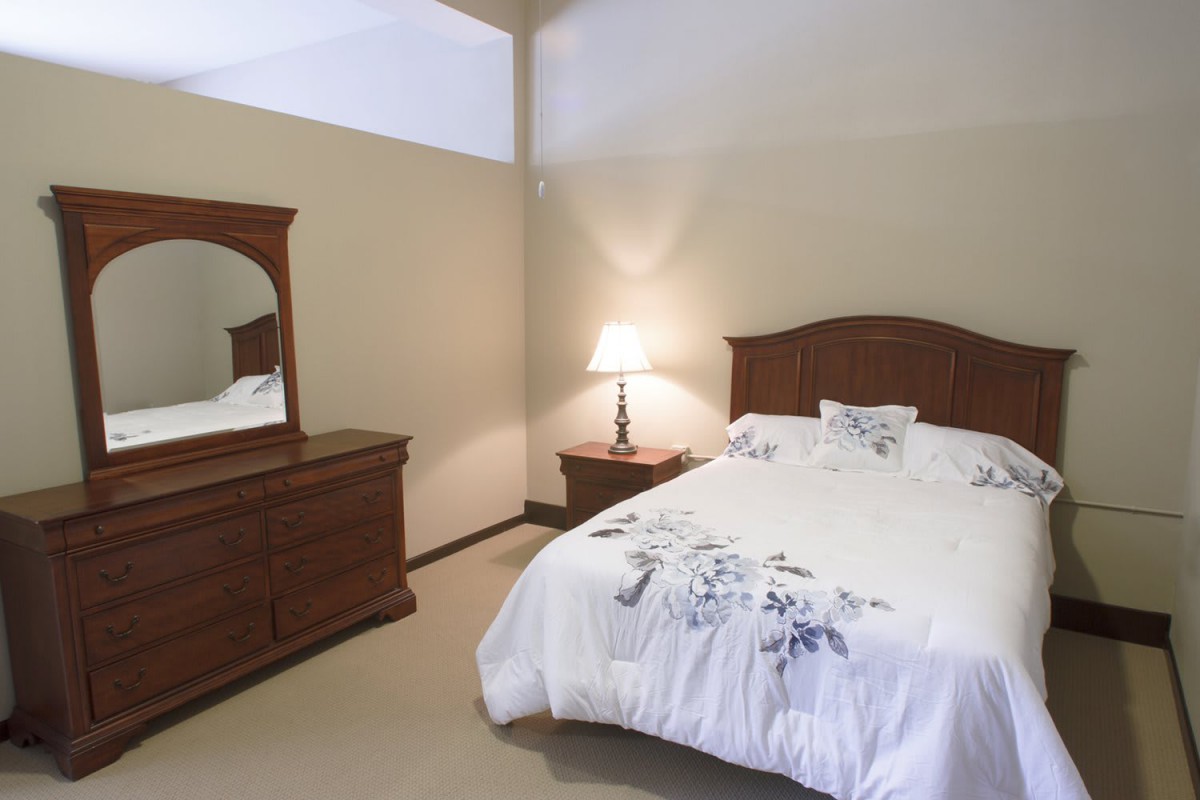 A cozy bedroom with a wooden bed, dresser, and nightstand, lit by a table lamp.