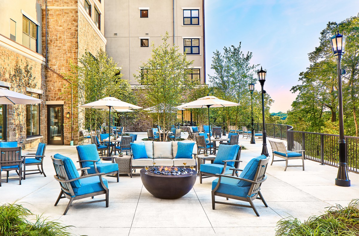 A modern outdoor patio featuring blue cushioned chairs, umbrellas, and a central fire pit in an urban environment.