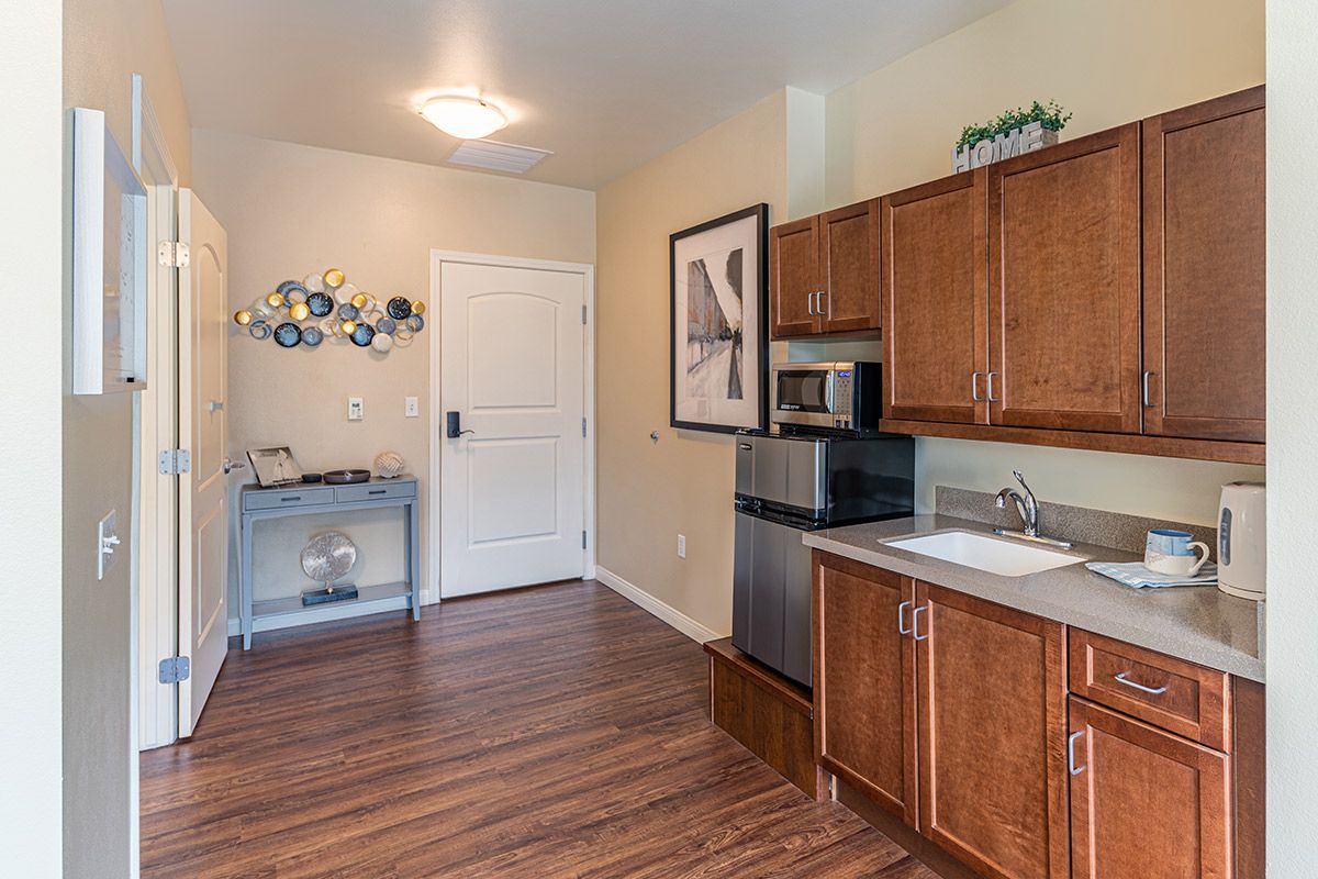 A small kitchenette with wooden cabinets, a microwave, and sink, featuring wall art and a hardwood floor.