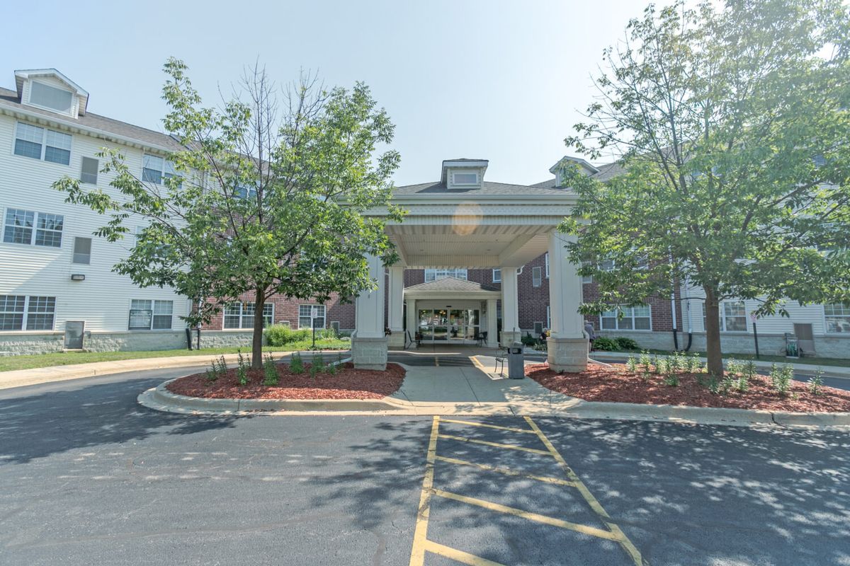 A building entrance with a covered drop-off area, surrounded by trees and landscaping, adjacent to a parking lot.