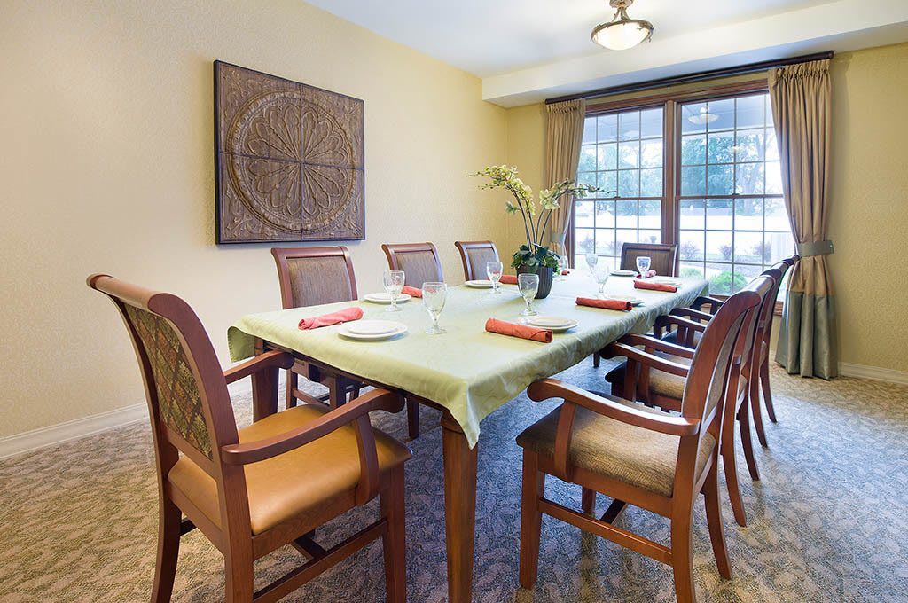 A neatly arranged dining room featuring a long table with chairs, decorated with a green tablecloth and floral centerpiece. Large windows provide natural light, and artwork adorns the wall.
