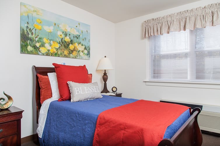 A cozy bedroom with colorful bedding, featuring a blue and red quilt, decorative pillows, and wall art above the bed.