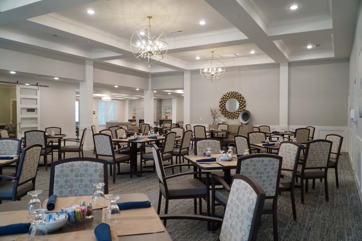An elegantly decorated dining room with multiple tables and chairs, featuring chandeliers and a mirror on the wall.
