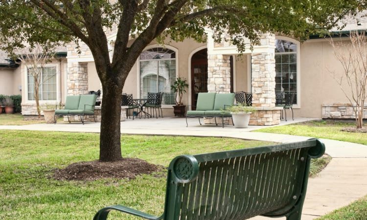 A peaceful outdoor seating area with a green bench under a tree, adjacent to a patio with chairs.