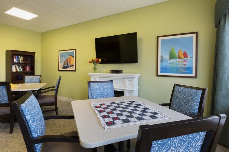 A cozy room with a chess board on the table, surrounded by chairs and a TV mounted on a light green wall adorned with colorful artwork.