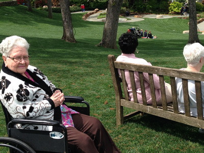 Two elderly women enjoying a sunny day in the park, one seated in a wheelchair and the other on a bench.