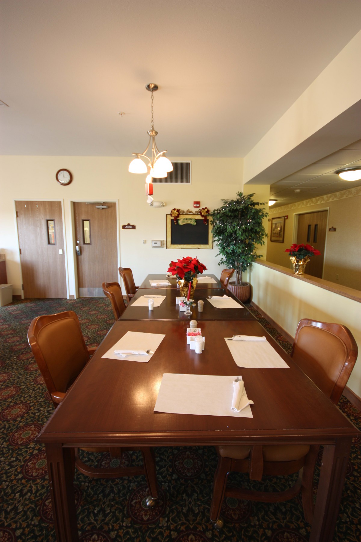 A neatly arranged dining room with a long wooden table, surrounded by chairs and set with placemats, featuring a centerpiece of flowers under warm lighting.