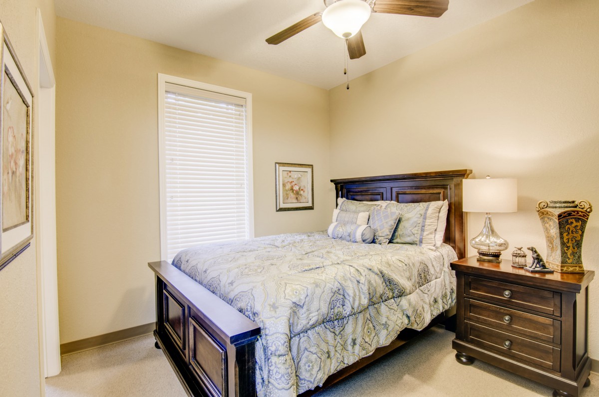 A cozy bedroom with a wooden bed frame, complemented by a matching nightstand and lamp, featuring a large window and ceiling fan.