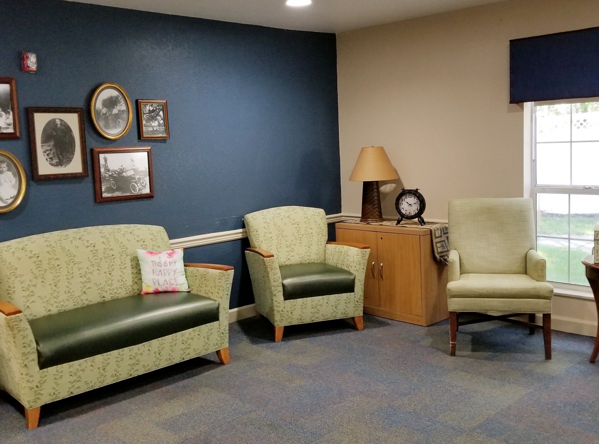 A cozy waiting room with green chairs, decorated with framed pictures and a lamp.