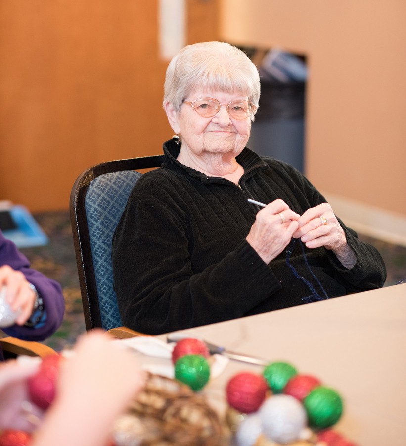 An elderly woman engaged in knitting at a table, surrounded by colorful yarn.