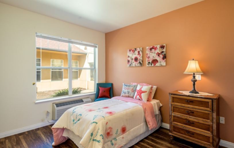 A cozy bedroom with a single bed featuring floral bedding, complemented by wall art and a warm-toned lamp.