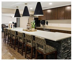 A modern kitchen featuring a long countertop with bar stools, pendant lighting, and dark wood cabinetry.