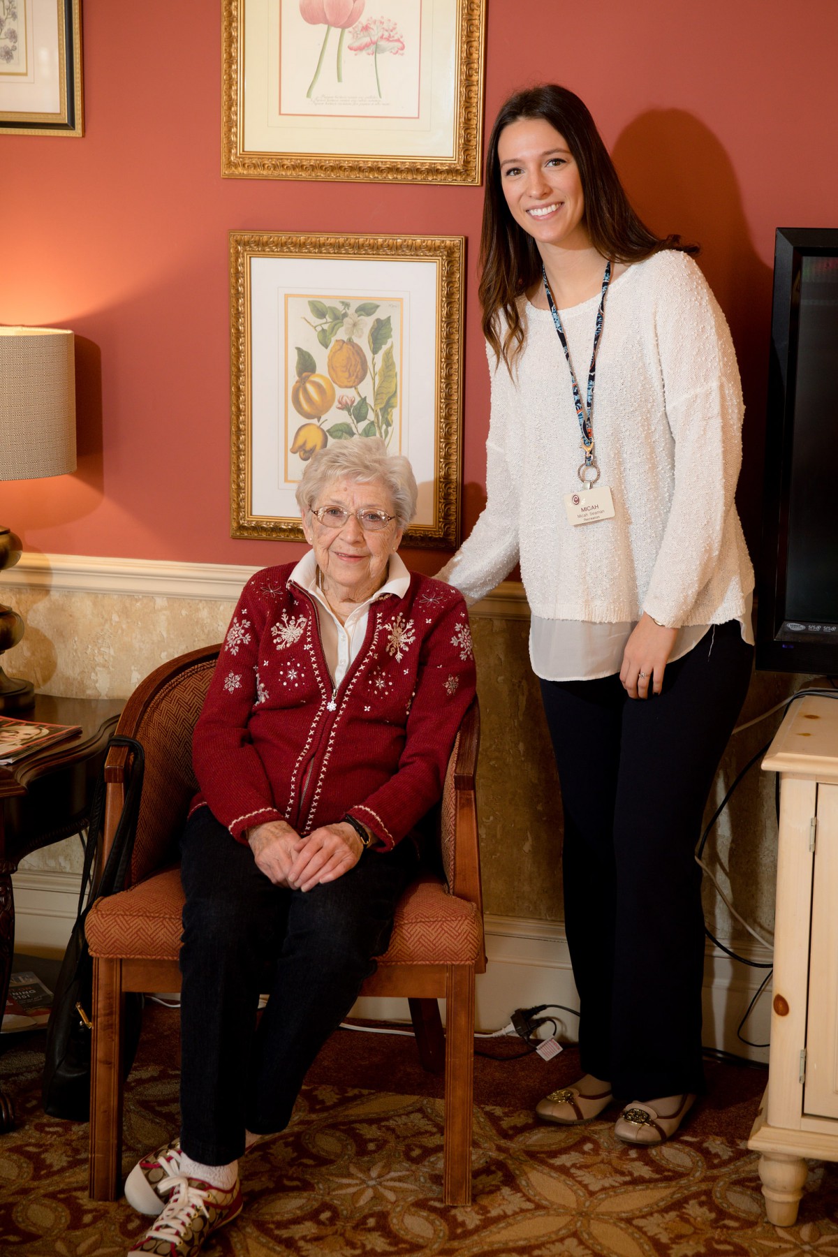 An elderly woman is seated in a chair while a young woman stands beside her in a living room setting.