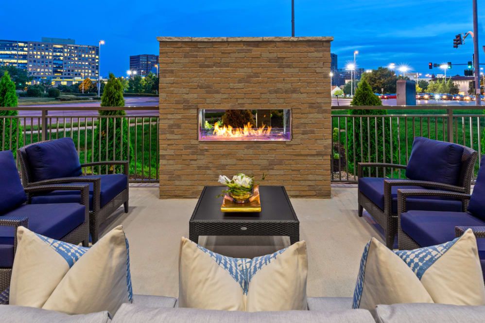 A cozy outdoor patio with a modern fireplace and comfortable seating, set against an evening cityscape.