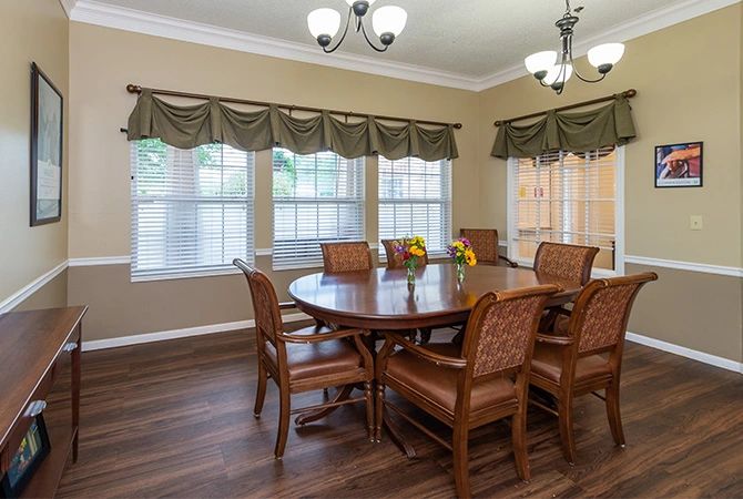 A spacious dining room featuring a large wooden table, surrounded by chairs, with windows adorned with curtains and two chandeliers providing lighting.