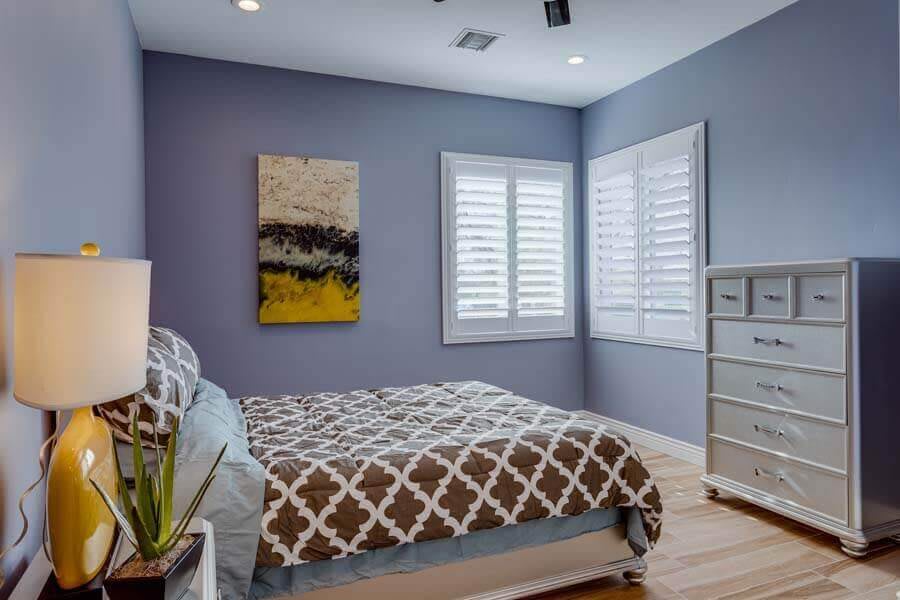 A modern bedroom with a patterned bedspread, abstract artwork on the wall, and white shutters on the windows.