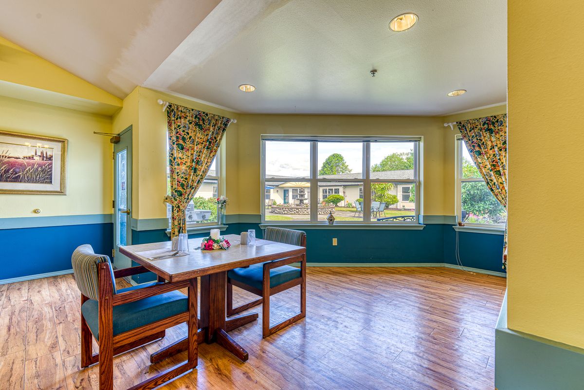 A cozy dining room with a wooden table and chairs by a large window, showcasing bright daylight and decorative curtains.