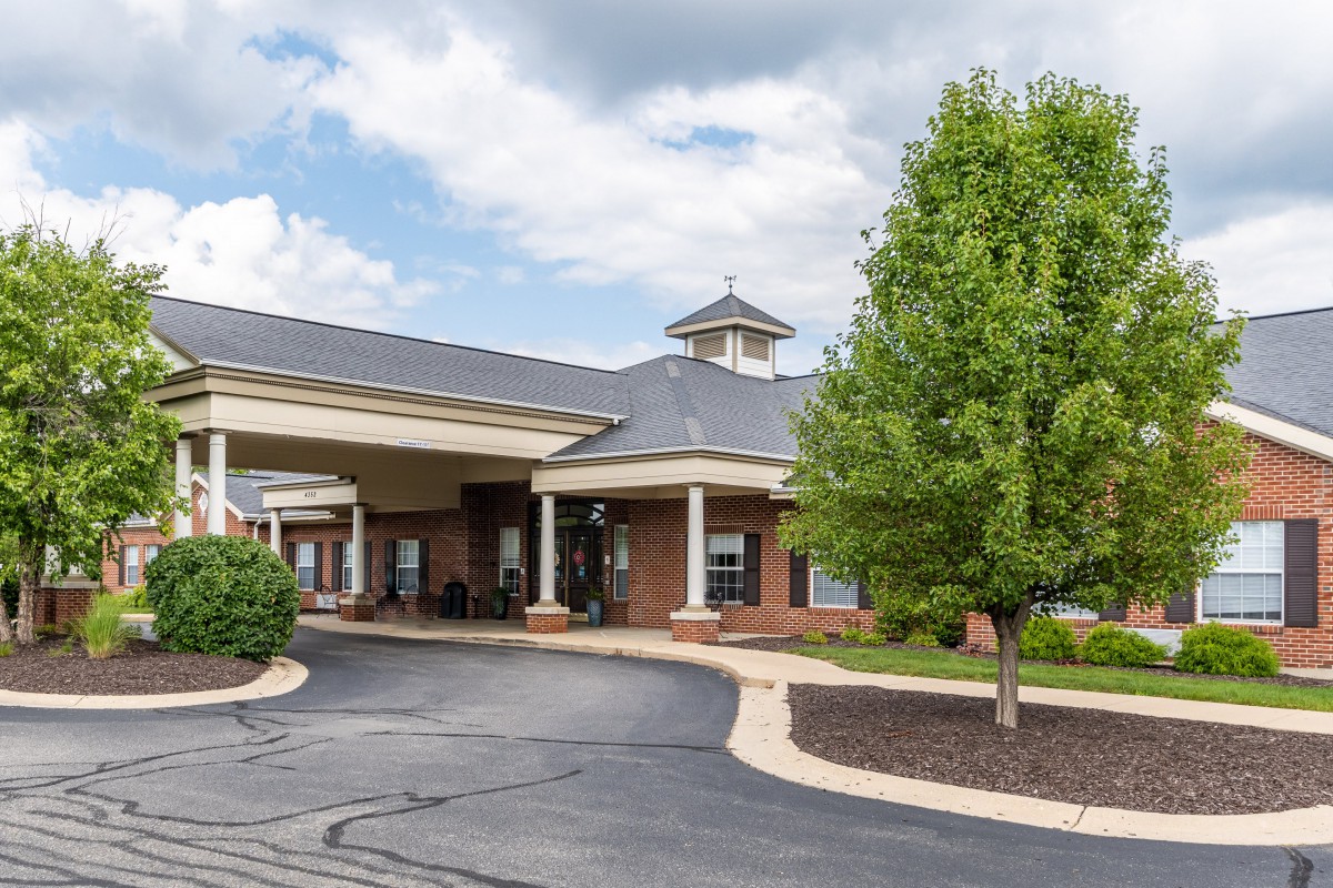 A brick building with a covered entrance and surrounding greenery under a cloudy sky.