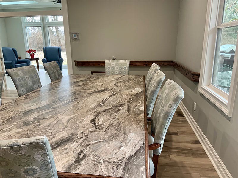 A spacious dining room with a large marble-patterned table and cushioned chairs, illuminated by natural light from a window.