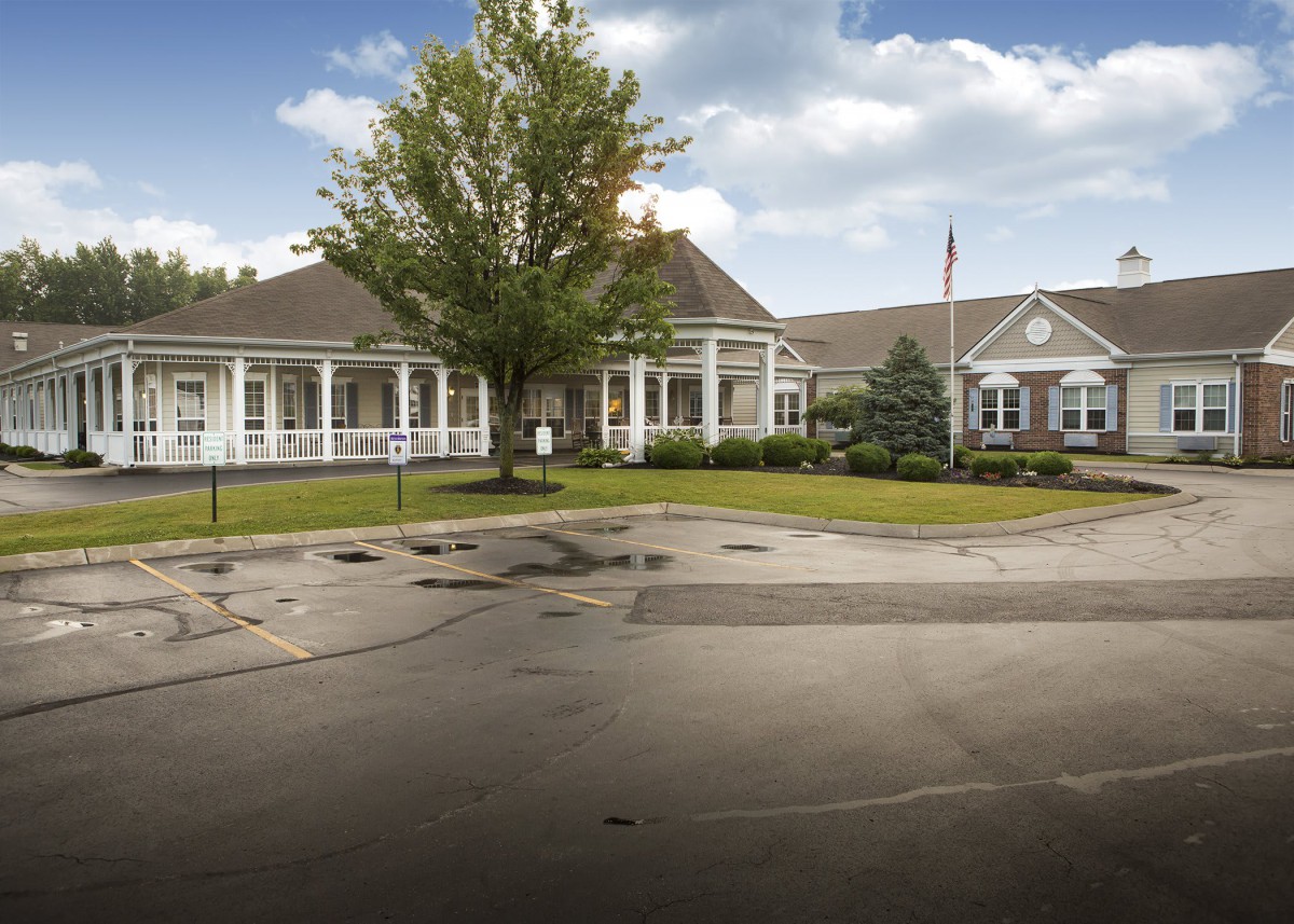 A large, single-story school building with a driveway and a tree in the foreground, set against a cloudy sky.