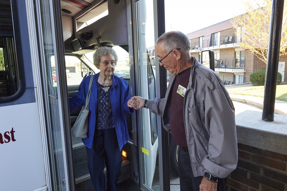 An elderly woman boards a bus assisted by an elderly man.