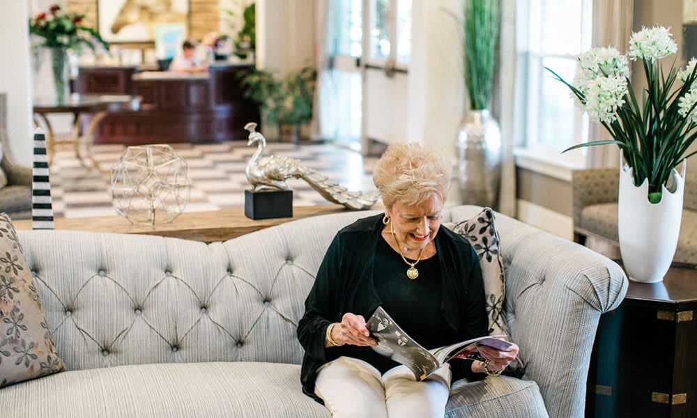 An elderly woman sits comfortably on a sofa while reading a magazine in a bright, inviting lounge area.