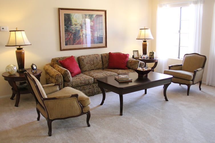 A cozy living room featuring a patterned sofa with red throw pillows, a matching armchair, and a dark wood coffee table. A lamp and wall art enhance the inviting atmosphere.