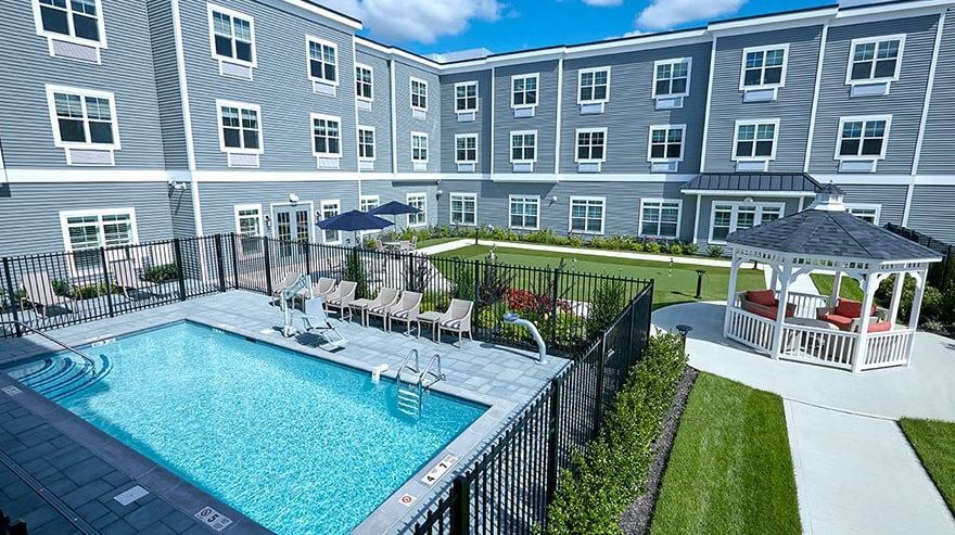 A modern apartment complex features a fenced swimming pool and well-maintained outdoor area under a clear blue sky.
