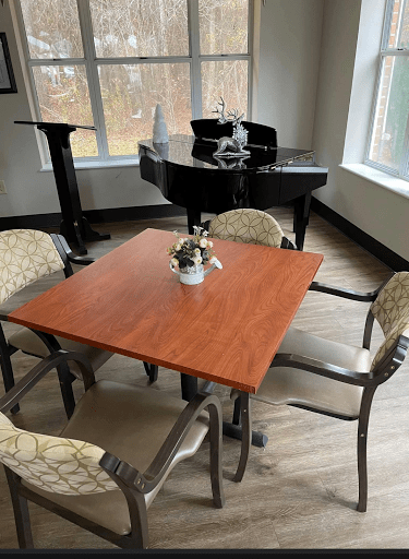 A cozy room with a wooden table surrounded by chairs, featuring a piano in the background and large windows letting in natural light.