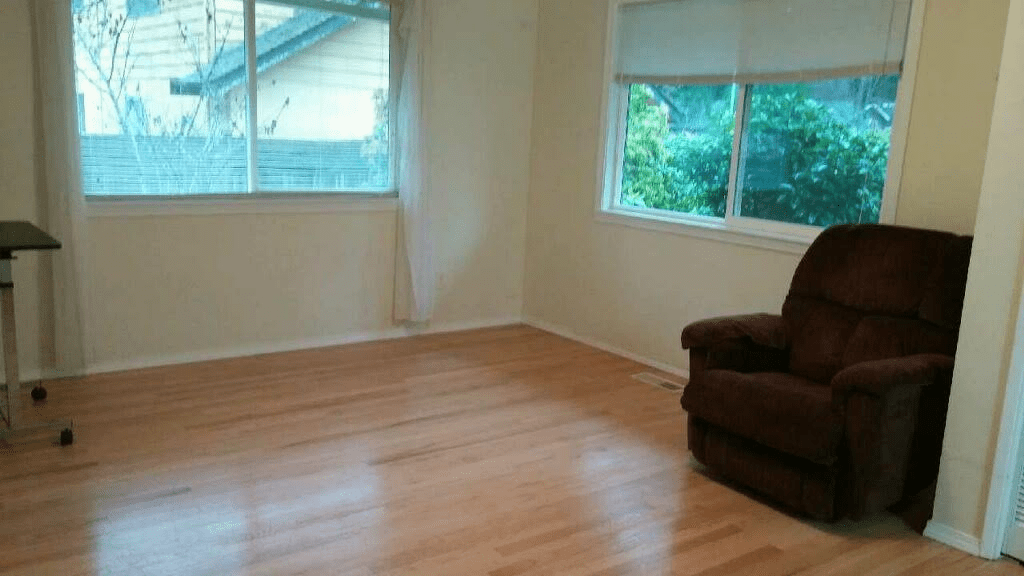 An empty living room with hardwood floors, a single armchair, and large windows letting in natural light.