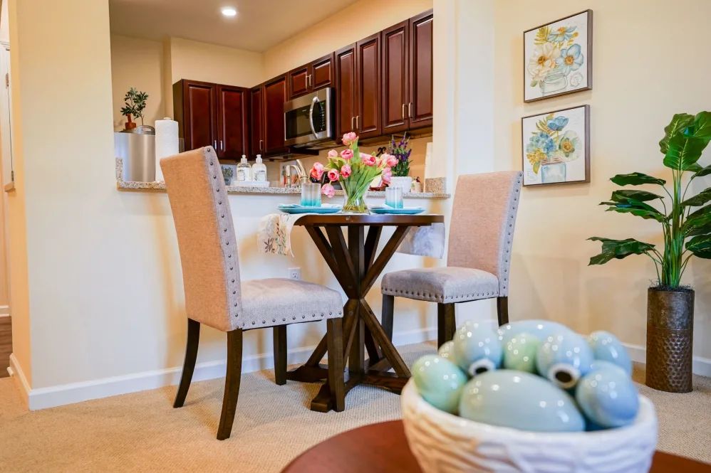 A cozy dining area with a small table and chairs, adorned with flowers and stylish decor, adjacent to a modern kitchen.