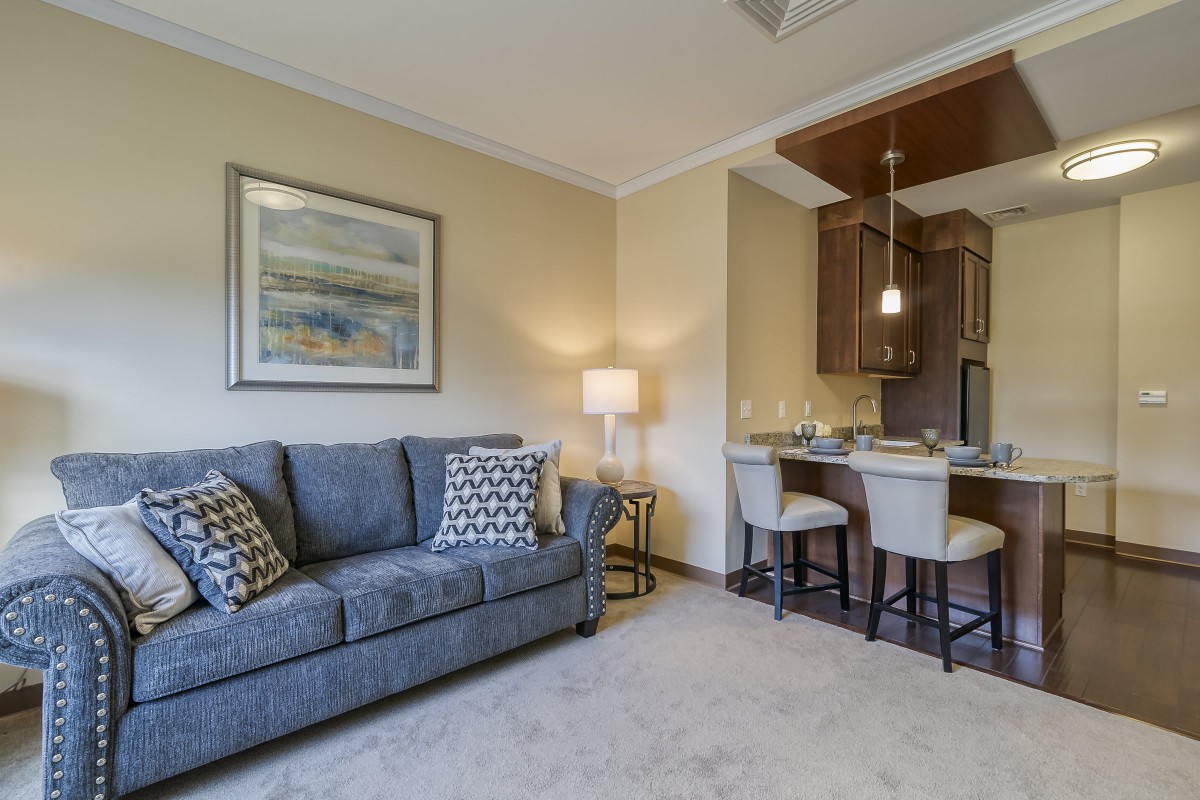 A cozy living room with a sofa and artwork, adjacent to a modern kitchen area with bar stools.